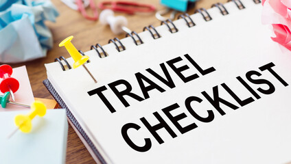 TRAVEL CHECKLIST - text on a notepad with wrinkled paper and paper needles on wooden background.