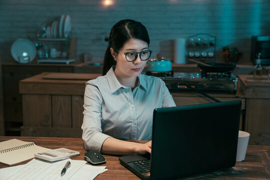 Attractive Young Asian Chinese Woman Working In Home Kitchen At Night. Office Lady Using Laptop Computer In Dark Workplace In Midnight. Concentrated Female Worker In Glasses Using Notebook Pc.