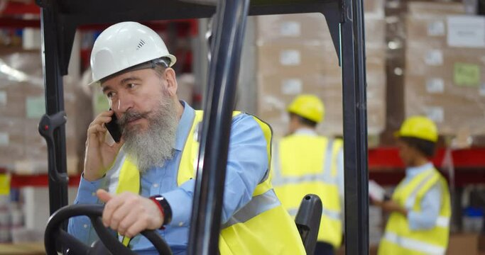 Senior forklift driver sitting in vehicle in warehouse talking on smartphone
