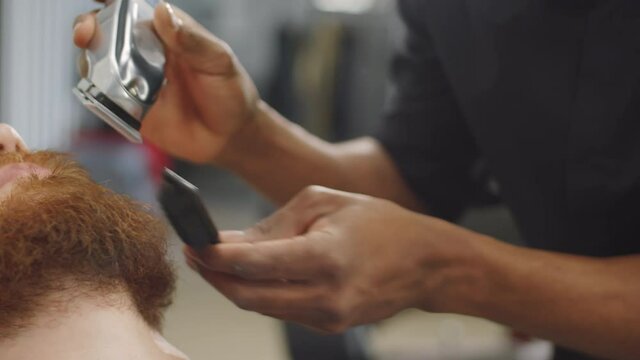 Tilt down close up shot of young Afro-American barber trimming beard of Caucasian male client with hair clippers while working in barbershop