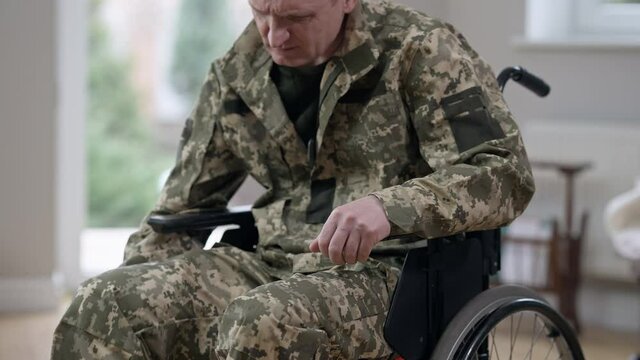 Serious Caucasian man in military uniform trying to stand up from wheelchair. Injured disabled middle aged soldier or veteran recovering at home indoors. Medical problems and lifestyle