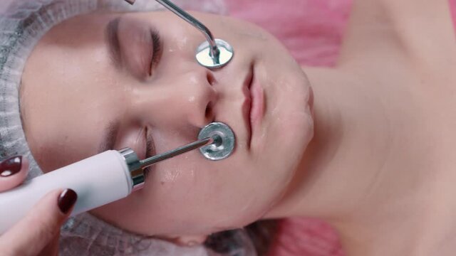 The beautician performs the procedure of microcurrent therapy of the facial skin. Close-up of the face, top view.