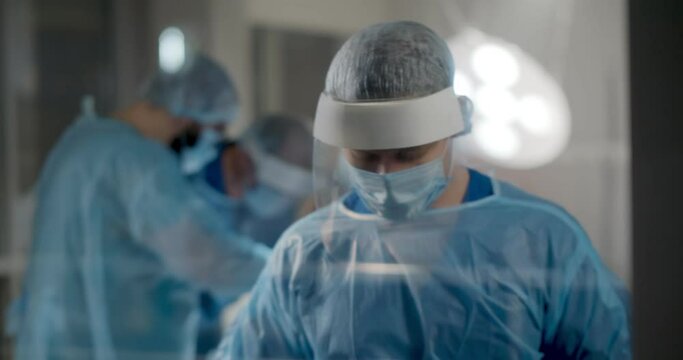 Portrait Of Male Surgeon Standing Near Glass Door While Team Performing Surgery In Operation Room