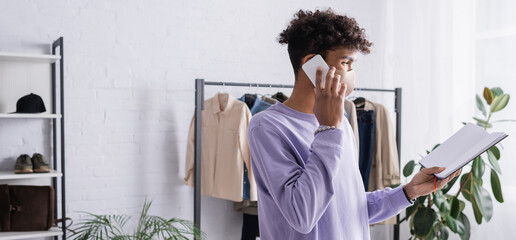 African american owner of showroom in protective mask talking on smartphone and holding notebook, banner