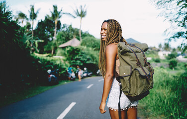 Joyful African American female tourist with touristic backpack smiling during summer vacations for visiting tropical island, happy dark skinned woman with travel rucksack exploring environment