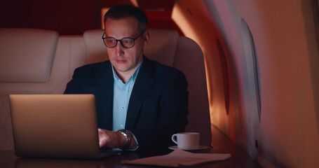 Successful businessman in glasses working on laptop sitting in chair of private jet.
