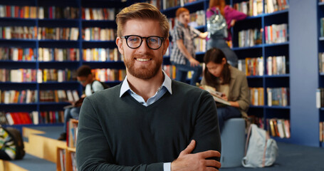 Portrait of young male teacher n library with other students studying in background