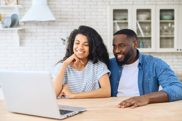 Happy African-American couple spends leisure time with the laptop in the modern kitchen at home, sits together at the countertop and watching movies, talking online with friends via video call