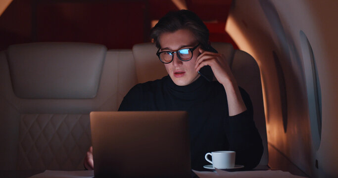 Young businessman using laptop and talking on phone traveling in private jet - Powered by Adobe