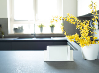 View of modern style kitchen. Mockup of a tablet with white blank screen over the kitchen table with yellow flowers. In the background, window illuminated by natural sunlight. Italian interior design