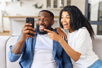 No way. Surprised multiracial couple looks at the smartphone screen with astonishment, African guy holds phone, charming biracial girlfriend points finger at screen, watching funny movies and laughing