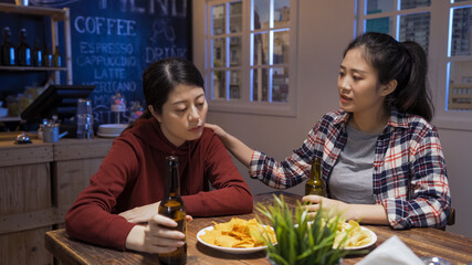 two depressed asian chinese female friends in night bar. Supporting sad friend lifestyle concept. group of young ladies talking and comforting with beer and snacks on wooden table in pub indoors.