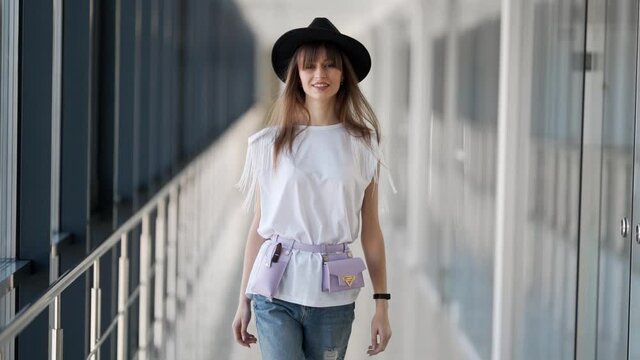 Beautiful girl in a hat with a waist microbag model gait walking through the glass corridors