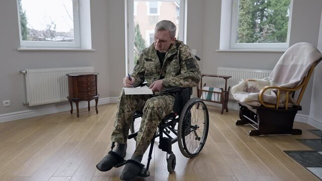 Wide shot portrait of concentrated paraplegic man in military uniform sitting on wheelchair writing letter. Focused handicapped disabled Caucasian veteran indoors at home. Lifestyle and disability
