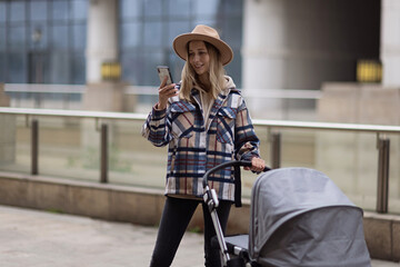 Happy Mother walking With Stroller In Park and using mobile phone. Joy of motherhood. Stylish young caucasian woman wearing warm clothes at autumn.