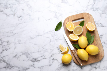 Many fresh ripe lemons with green leaves and knife on white marble table, flat lay. Space for text