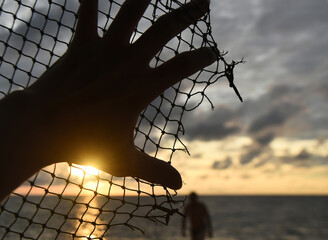 The hand lies on a rope net with a torn edge against the background of a sea sunset, the silhouette...