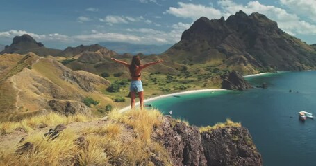 Young woman rising hands on mountain top above sea aerial. Summer nature landscape. Girl tourist standing on cliff at seaside. Lady admires tropic seascape with ships, boats. Active tourism lifestyle