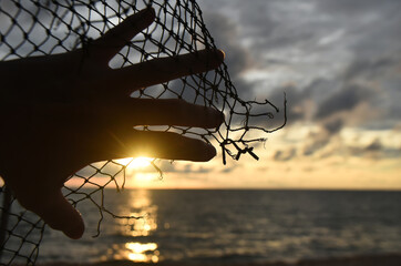 A hand lies on a rope net with a torn edge against the background of a sea sunset