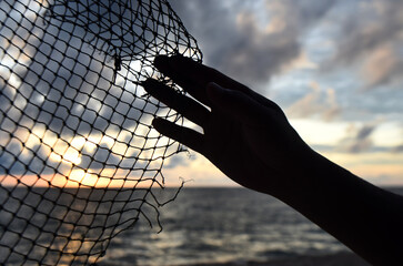 The hand holds a rope net with a torn edge against the background of sea sunset