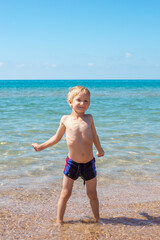 A little blond boy in swimming trunks plays on the seashore, stretches and smiles, standing in the water. Summer happy holidays.