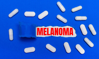 White pills lie on a beautiful blue background. In the center is white paper with the inscription MELANOMA. Medical concept. View from above.