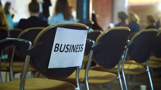 Word business printed on sheet of paper attached to chair with sticky tape, blurred females on background. Pan shot participants of business conference for women. Concept of success