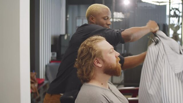 Cheerful Afro-American barber taking off haircutting cape, talking and shaking hands with bearded male client in barbershop