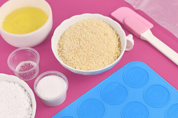 Ingredients for making homemade French Macaron sweets including powdered sugar, ground almonds, egg white, salt and mold mat on pink background