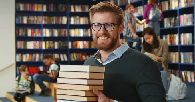 Bearded school teacher or librarian holding books in hands standing in library interior.