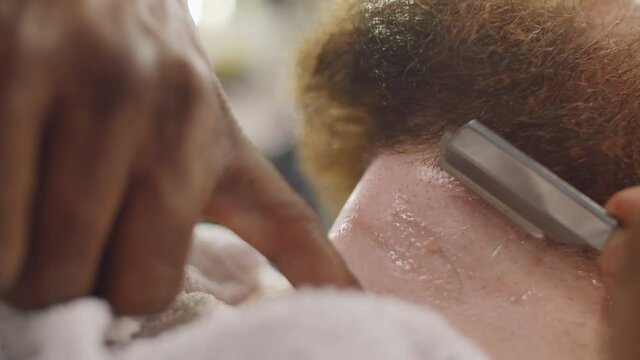 Close up view of hands of professional barber using straight edge razor while shaving neck of man in barbershop