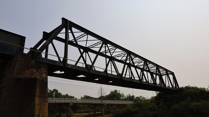 Old structure of the railway bridge and pier. Outdoor black metal frame bridge to cross the river from side view. On a concrete bridge background and clear sunny blue sky. Selective focus