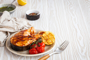 Grilled salmon steak glazed with teriyaki sauce, vegetables and lemon served on ceramic plate on rustic white wooden table from angle view, selective focus with space for text
