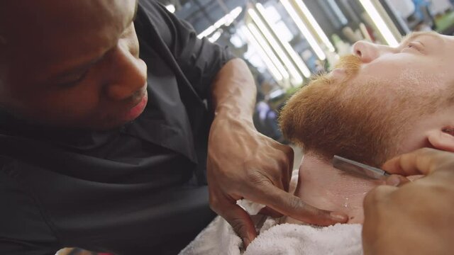 Professional Afro-American barber shaving neck of Caucasian bearded man with straight edge razor in barbershop