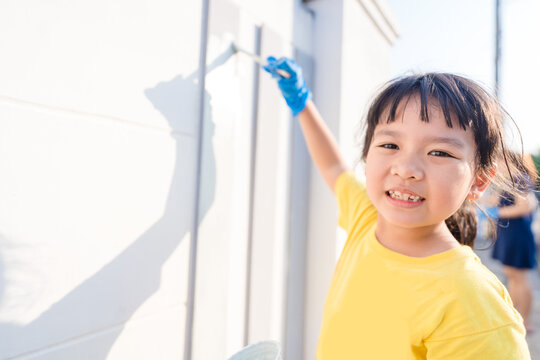 Cute little asian kid girl painting on wall cement fence outdoor.Volunteer kid project painting wall in local school.renovate.kid holding brush.renew house.Paint color.Home, community service.charity.