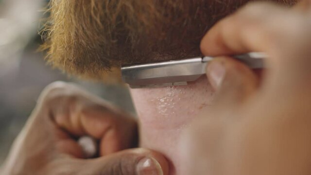 Close up shot of hands of unrecognizable male barber shaving beard of client with straight edge razor in barbershop