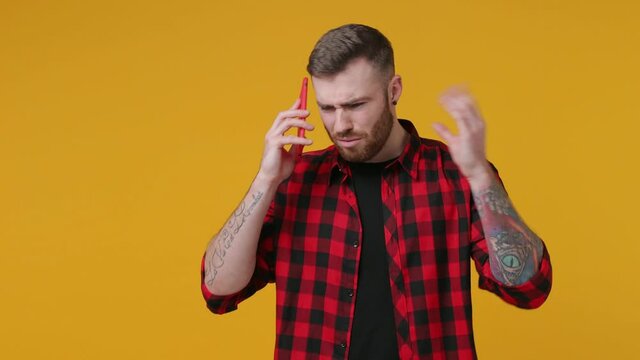 Irritated tattooed bearded young man 20s in red shirt isolated on yellow background studio. People lifestyle concept. Talking on mobile cell phone swearing showing blah blah gesture ja jaja hands