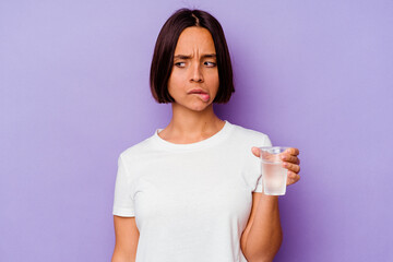 Young mixed race holding a glass of water isolated on purple background confused, feels doubtful...