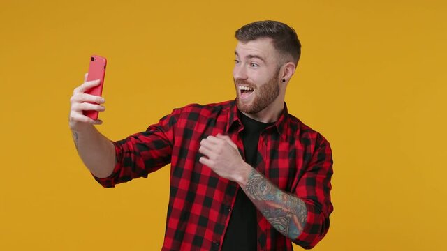 Cheerful tattooed bearded young man 20s in red shirt isolated on yellow background studio. People lifestyle concept. Doing selfie shot on mobile phone showing thumb up biceps muscles ok okay gesture