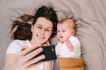 Top view of sweet baby girl with happy young caucasian mother take make selfie on phone, lying together on bed. New generation, gadgets easy usage in everyday life, entertaining having fun concept