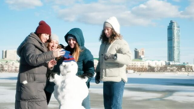 four girls in the city are making a snowman. Taking pictures. Selfie. Long time no see. They fool around, have fun on a snowy day in the park. Friendship concept. Stroll. Winter.