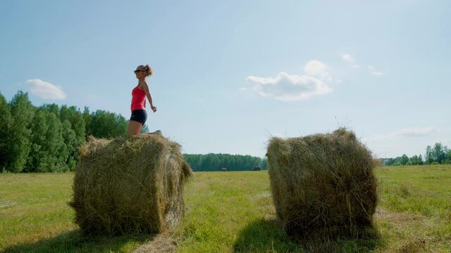 Elegant girl in the summer in the field jumps from haystack to haystack. Beautiful girl on a sunny day in shorts, red T-shirt and glasses jumps on the straw hay.