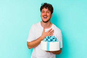 Young caucasian man holding a cake isolated on blue background laughs out loudly keeping hand on chest. - obrazy, fototapety, plakaty