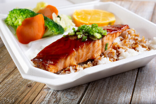 A view of a to-go container featuring teriyaki salmon and rice as a combination plate.