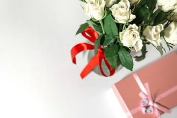 White tea roses stand in a glass vase, which is tied with a red ribbon and stands on the table. There is a gift nearby. in a pink box. Gift bouquet