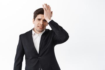 Worried and troubled corporate man hold hand on forehead and frowning upset, have problem, remember forget, standing over white background in black suit