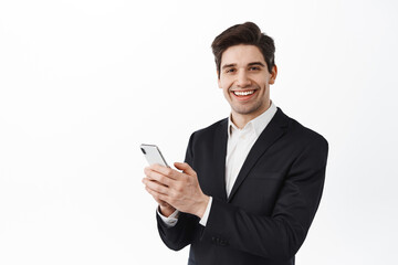 Corporate man, businessman in black suit using mobile phone, standing with smartphone and looking at camera, smiling, white background