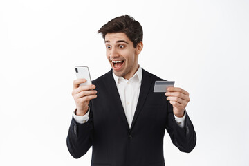 Excited businessman holding credit card, looking happy at smartphone screen, receive money transfer, standing over white background