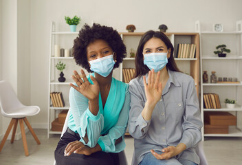 Two female colleagues, coworkers, modern vloggers, presenters or friends wearing masks waving hello...