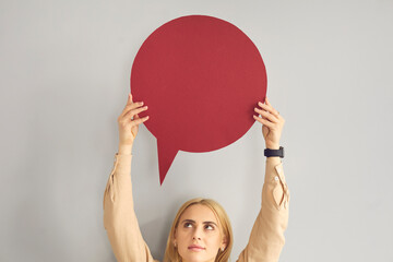 Happy young woman or teen girl holding up one dark wine red round clean free mockup copyspace speech bubble on gray background. Mock up balloon for message notification or expressing people's opinion